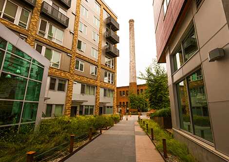 A narrow pathway lined with greenery runs between modern apartment buildings, leading to a tall, narrow brick chimney in the background. The facades feature large windows and mixed materials.