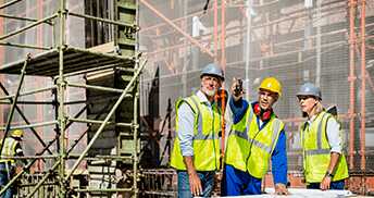 Three construction workers wearing safety vests and hard hats stand near scaffolding on a building site. One worker points upwards, discussing plans, while the others listen attentively.