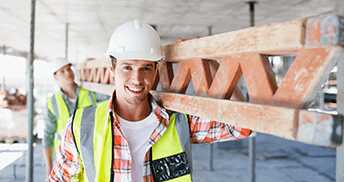 Two construction workers in safety gear and helmets are on site. The front worker smiles while carrying a metal beam on his shoulder. The second worker is slightly out of focus in the background.