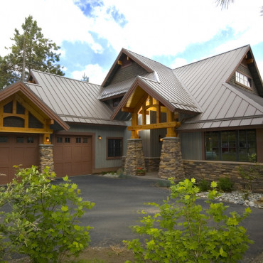 A rustic house with a steep metal roof, large wooden beams, and stone accents. It features a two-car garage, a paved driveway, and is surrounded by green trees and shrubs under a partly cloudy sky.