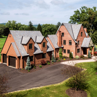A large red brick house with multiple gables, surrounded by a paved driveway and landscaped gardens. The home features a two-car garage and is set against a backdrop of trees and a lush green lawn under a partly cloudy sky.