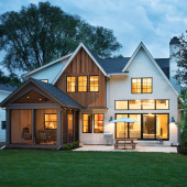A modern two-story house with large windows illuminated at dusk. The exterior features a combination of white and wood siding. A patio with a table and umbrella is in the backyard, surrounded by a well-kept lawn and trees.
