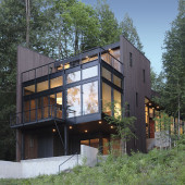 A modern, two-story house with large glass windows and dark exterior stands amidst lush greenery. The house features an angular design with a concrete base, surrounded by tall trees under a clear blue sky.