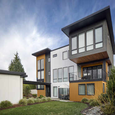 Modern two-story house with a mix of gray, white, and yellow-brown exterior panels. Features large windows, a balcony, and a covered patio area. A small lawn and plants are in the foreground, with a partly cloudy sky overhead.