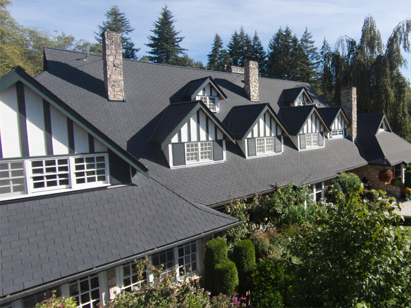 A large PNW Tudor-style house with a dark shingled roof, multiple dormer windows, and several tall stone chimneys.