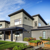 Modern two-story house with a combination of light green and gray siding. It features large windows, a sloped roof, and a manicured garden with rectangular wooden planters in front. The sky is clear and blue.