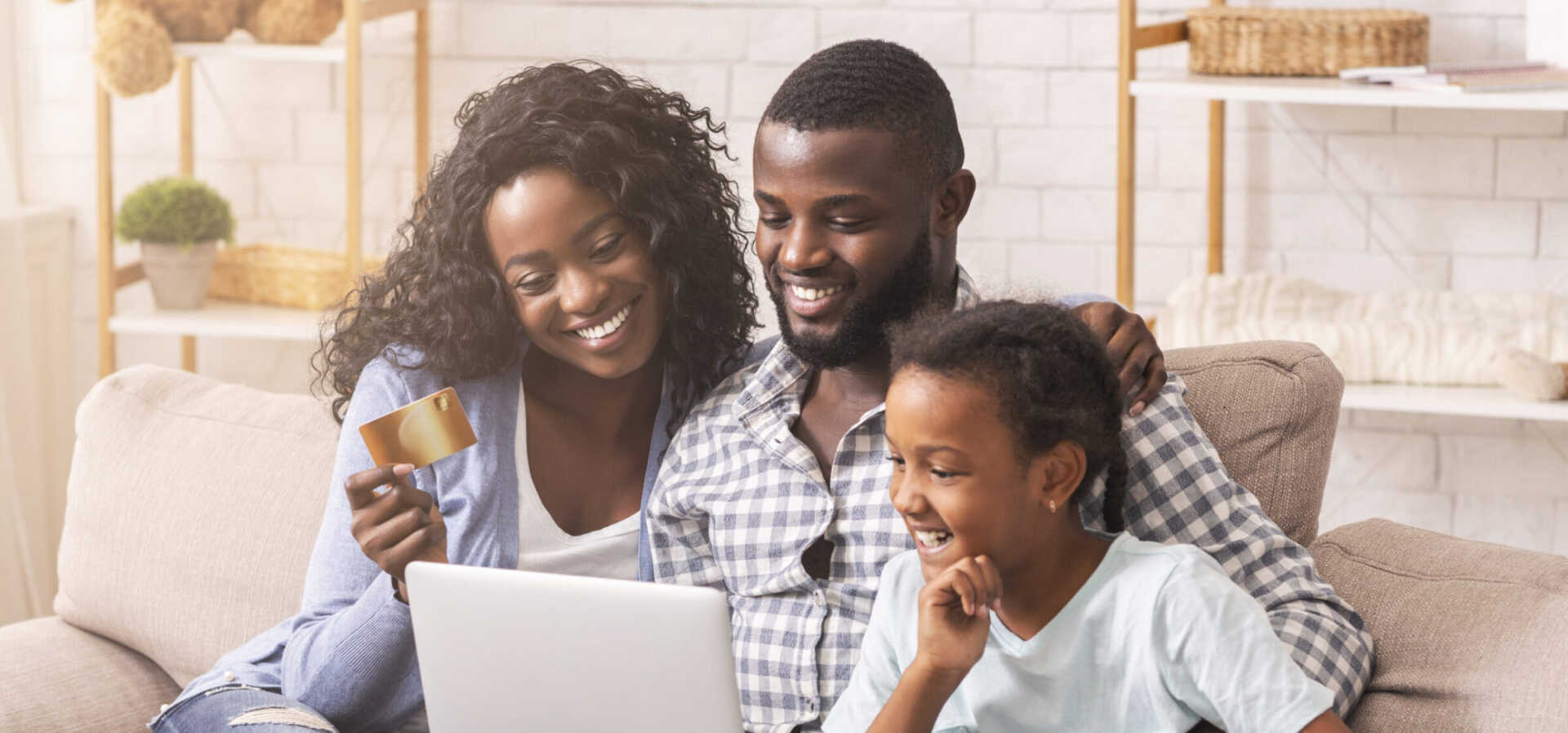 A smiling family sits on a couch, looking at a laptop. The woman holds a credit card, while the man and a child are engaged with the screen. The room is bright and warmly decorated.
