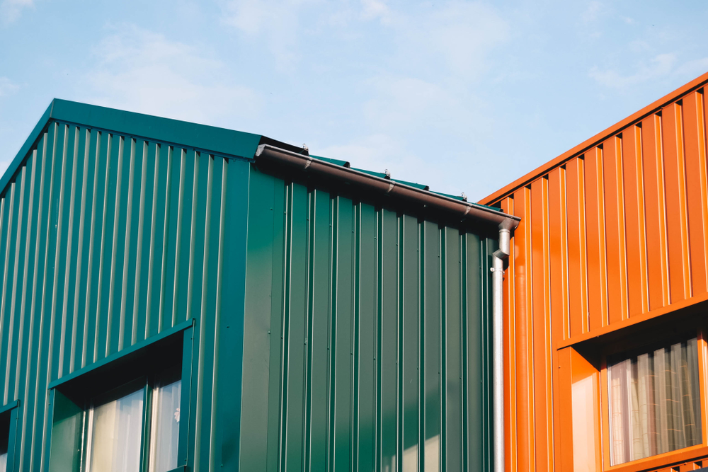 Two modern buildings with corrugated metal exteriors against a blue sky. The left building is dark green, and the right one is bright orange. Both have large windows reflecting the sky.