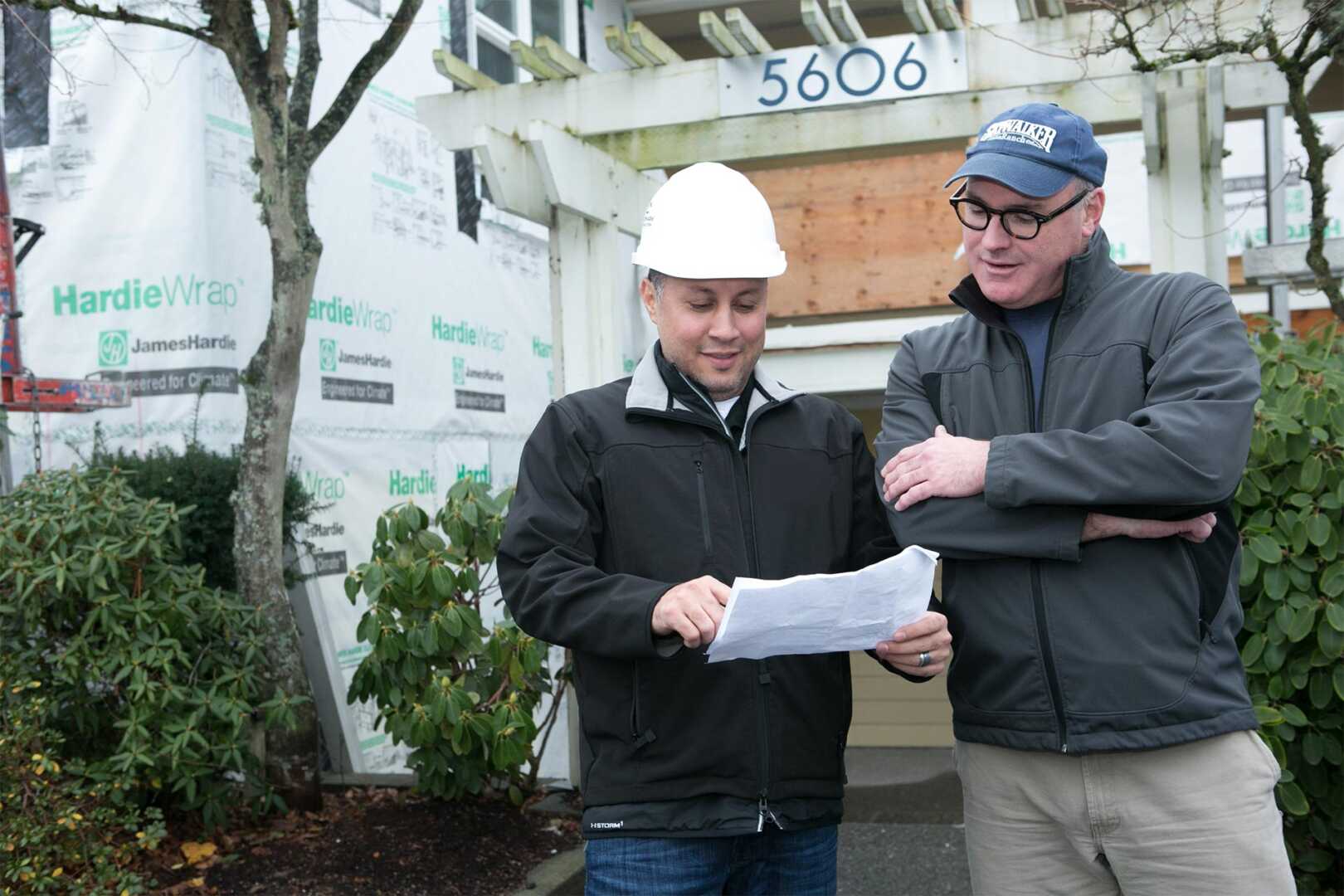 Two men in jackets stand outside a house under construction. One wears a white hard hat and holds a paper, while the other wears a cap. They appear to be discussing building plans, with part of the house wrapped in construction material in the background.