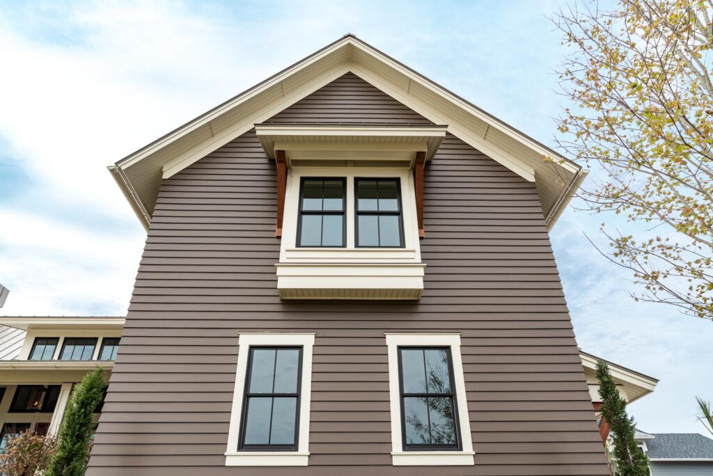 A brown house with white trim is shown from a low angle. It has two stories with three windows visible on the side, and the sky is partly cloudy. Theres a tree with sparse leaves on the right.