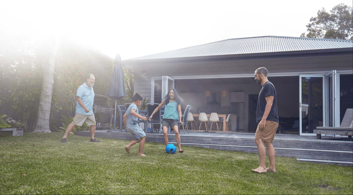 A family of four playing soccer on a grassy lawn in front of a modern house. Two adults and two kids are engaged in the game, with the houses sliding glass doors open, revealing indoor seating. The sky is bright and clear.
