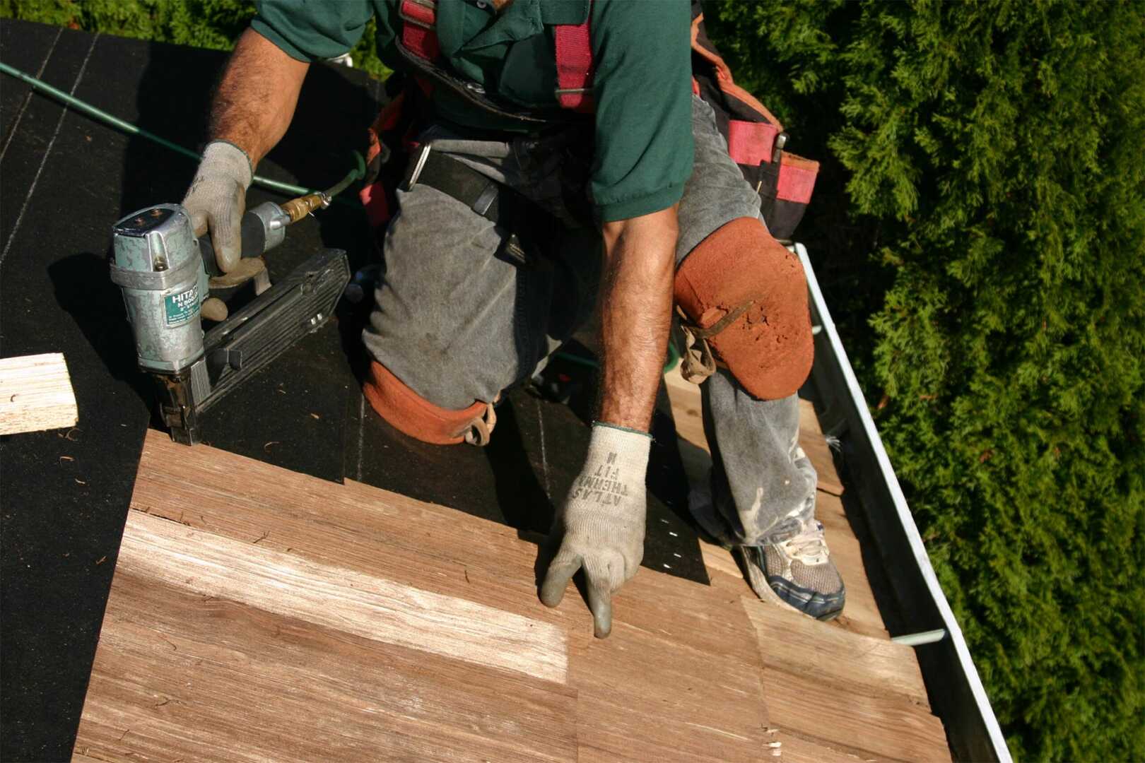 A construction worker kneels on a roof, using a nail gun to secure wooden shingles. They wear knee pads, gloves, and a safety harness. Parts of the roof are lined with black underlayment. Green trees are visible in the background.