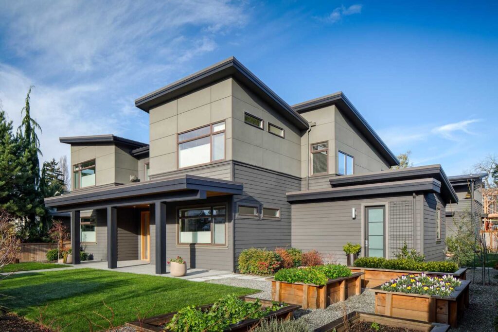 Modern two-story house with gray siding and large windows, surrounded by a landscaped yard. The front features a wooden door, manicured lawn, and raised garden beds filled with flowers. Bright blue sky and light clouds above.