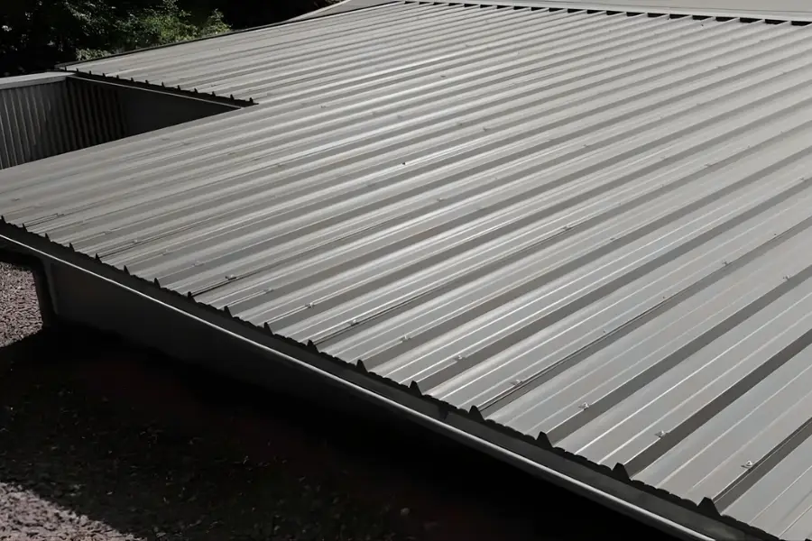 A close up view of a gray corrugated metal roof with visible screws and ridges, taken from an elevated angle under daylight. Trees and gravel are partially visible around the edges.