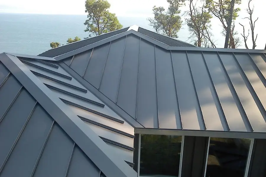 A modern metal roof with standing seam panels, featuring clean lines and sharp angles, overlooking a body of water and surrounded by tall trees in the background.