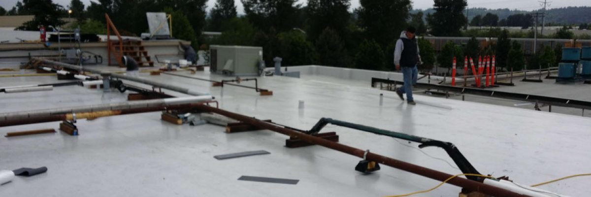 A person walking on a flat rooftop surrounded by equipment and materials. The sky is overcast, and there are trees and buildings in the background. Construction tools and pipes are scattered on the roof.