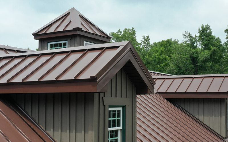 Brown metal roofing with visible ridges covers a house, featuring dormer windows and a cupola. The roof contrasts against a backdrop of green trees and a cloudy sky.