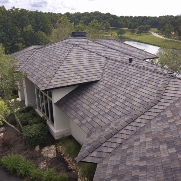 Aerial view of a large modern house with a complex, multi-faceted roof covered in gray shingles. The house is surrounded by lush greenery and is situated near a serene pond, with a clear sky overhead.