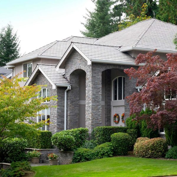 A two-story stone house with arched accents surrounded by lush greenery and trees. The front door has decorative wreaths, and there is a well-maintained lawn and shrubs in the foreground.