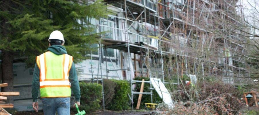 A construction worker wearing a hard hat and reflective vest walks toward a building covered in scaffolding. The surrounding area has trees and some construction materials scattered on the ground.