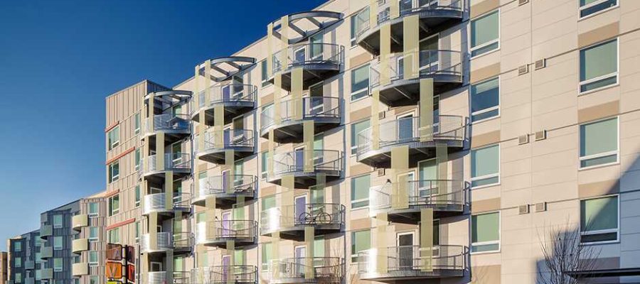 A modern apartment building with multiple floors featuring balconies and large windows. The exterior is a mix of light panels and glass. Clear blue sky in the background.