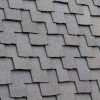 Close-up of gray, rectangular asphalt shingles on a roof, arranged in a staggered pattern. The texture is rough and gritty, with the shingles overlapping one another, creating a layered effect.