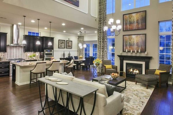 Spacious open concept living area with a modern kitchen, dining space, and living room featuring dark wood floors, high ceilings, large windows, a stone fireplace, and elegant decor with neutral tones and contemporary furniture.