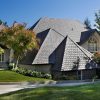 A large house with a gray tiled roof sits amidst lush greenery. It features multiple gables and large windows. The front yard is well-maintained with a curved driveway and various trees and shrubs surrounding the home.