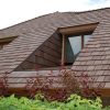 A house with a brown shingled roof featuring a triangular dormer window. The roof has wooden trims, and red-leaved shrubs are visible in the foreground. The sky is partially visible above the roof.