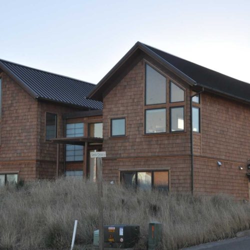 A two-story wooden house with a shingle exterior and multiple large windows sits on a slight hill. It has a dark metal roof and an attached garage. Tall grasses surround the building. A street sign is visible in the foreground.