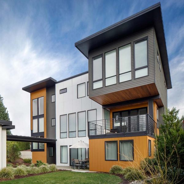 Modern multi-story house with a mix of white, black, and orange-brown panels. Large windows dominate the design. A small patio with a table and umbrella is surrounded by landscaping, set under a partly cloudy sky.